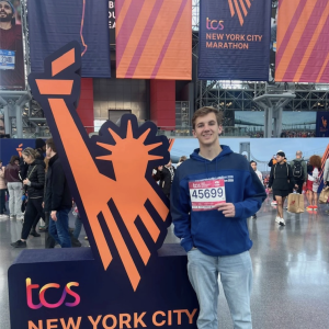Damon Holbrook standing in front of the TCS New York City Marathon sign, holding his race bib.