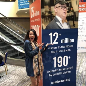 Kristin Schneider Smedley poses in front of a standing informational display featuring a photo of H. Eric Hartman, CRF Director of Advocacy, from RareDiseases.org. The sign notes that the National Organization for Rare Disorders (NORD) website had more than 12 million visitors in 2018 from over 190 countries.